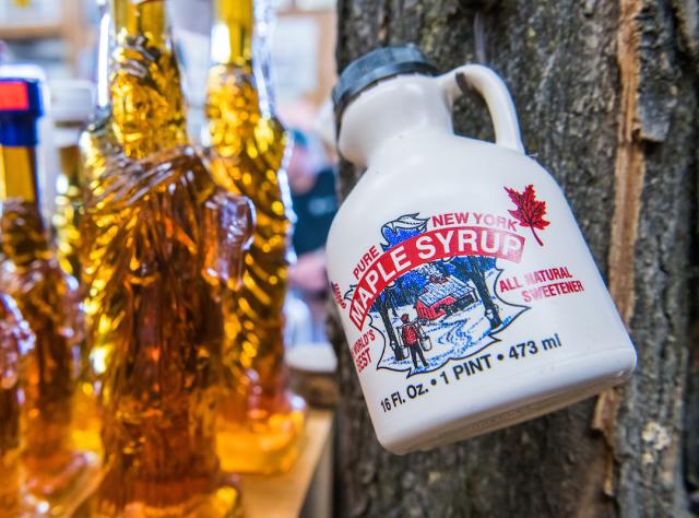 A bottle of New York Maple Syrup hangs on a tree with glass bottles of amber maple syrup sit nearby