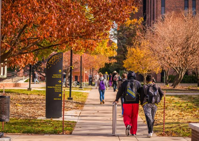 Students walk along a paved path lined with fall foliage and downed leaves at Wichita State University