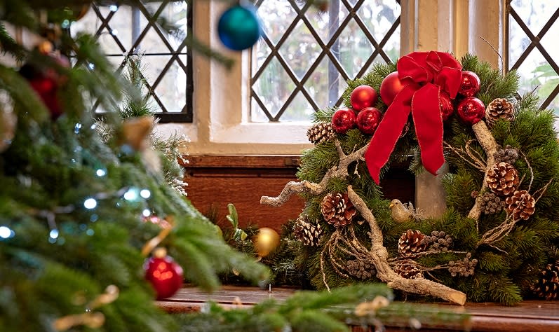 A Christmas wreath with bright red bow and pin cones sits on a windowsill by a Christmas tree