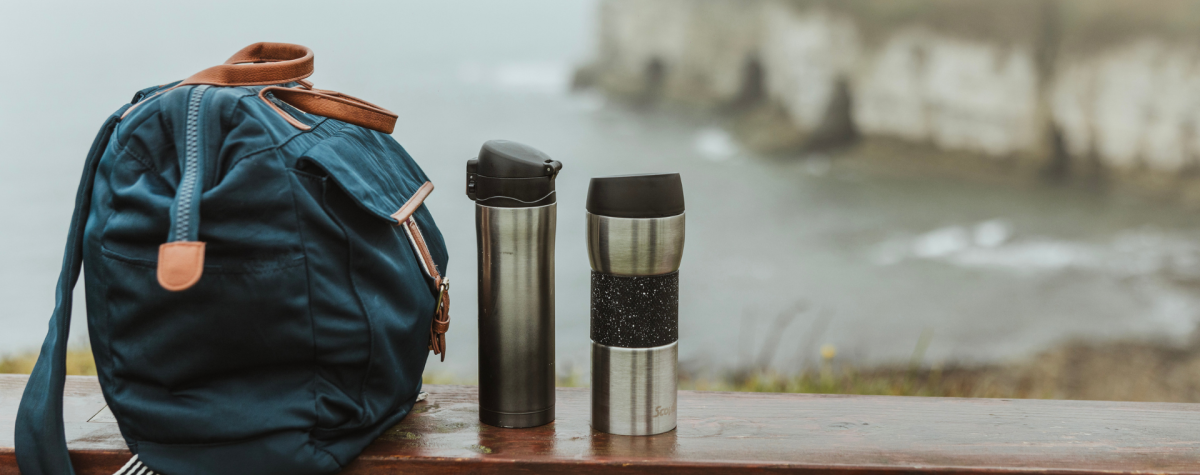 A set of two flasks sat on a bench alongside a backpack overlooking the sea with the cliffs in view to the right.