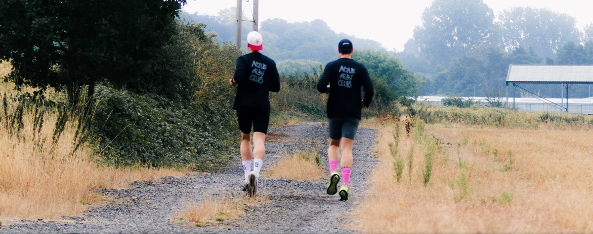 Two runners heading away down a gravel single track path, hedgerow to the left and field to the right.