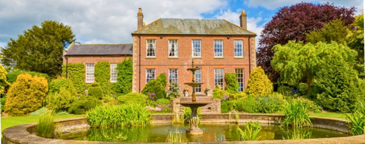 An external view of The Wold Cottage which shows the building surrounded by trees and a fountain to the front of it.