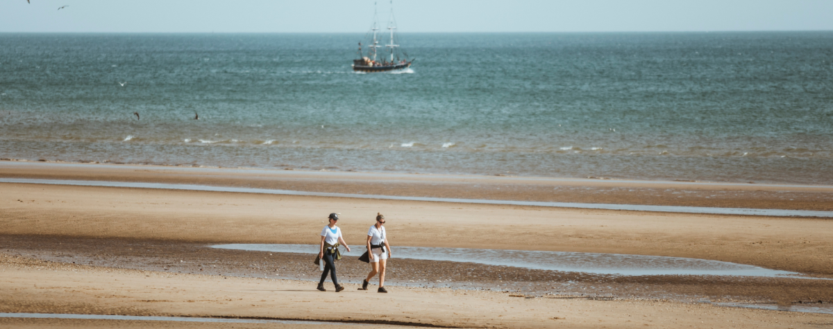 Two women walking along Bridlington's South Beach. A boat is visible in the sea behind them.