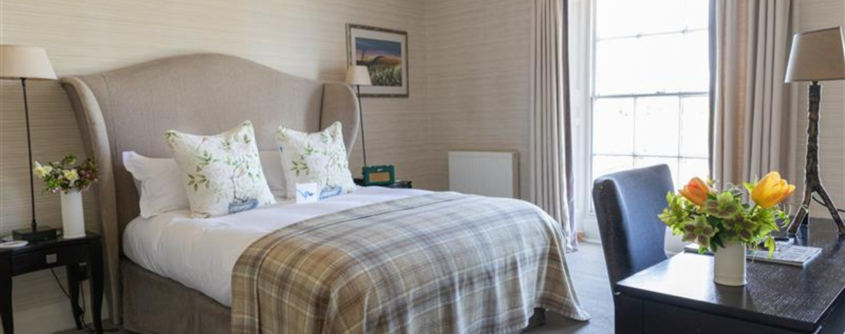 A bedroom at Tickton Grange Hotel near Beverley. The bed is dressed in neutral linens and two lamps sit on bedside tables at both sides. In front of the bed is a desk and chair and on the desk sits a case of orange flowers.