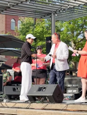 Ruben Mendoza (left, wearing a black polo, white slacks, and white ballcap) shakes hands with Nick Wolda (wearing a red T-shirt, white blazer, and navy slacks) as he accepts his 2025 NTTW Hospitality Award. They're standing on the stage at Market Street during the Spring Concert Series.