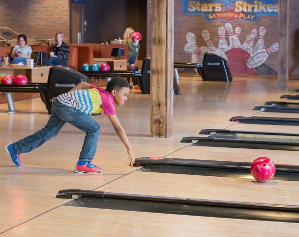 Child rolling a bowling ball at a bowling alley with a Stars and Strikes logo on the wall behind them