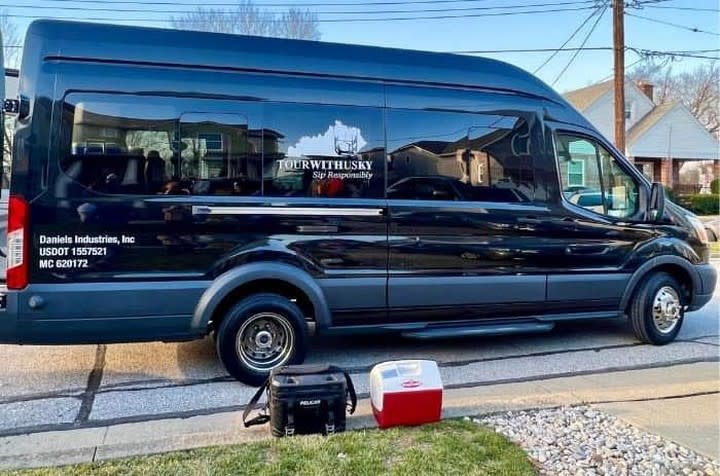 A black, passenger van sits on the street. In front of it are two small coolers. The image on the van says TourWithUsKy and has a picture of the state of Ky.