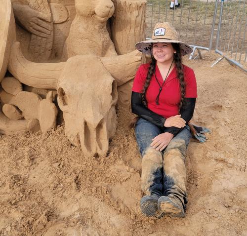A sand sculptor in a wide-brimmed hat with braids sits in the sand next to her sculpture. Only part of the sculpture is visible, but what the viewer can see is of a Western-style skull.
