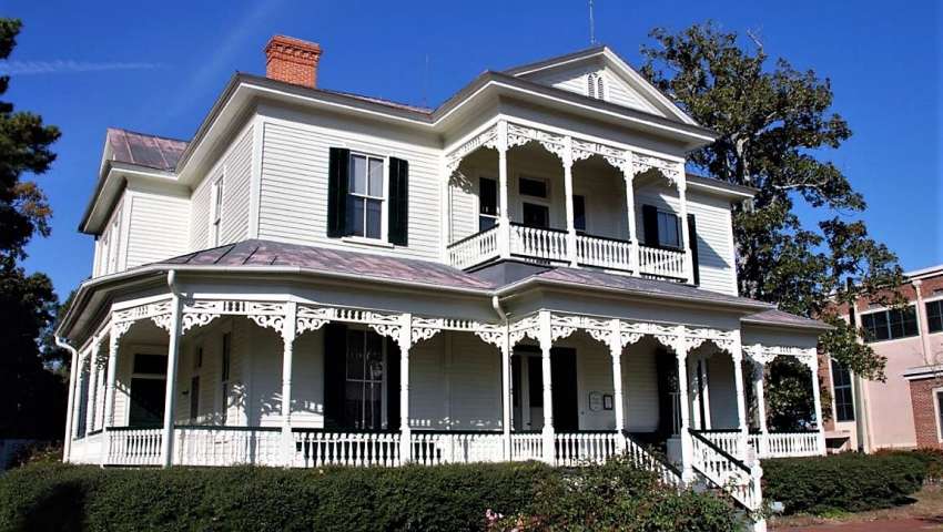 The 1897 Poe House, a two-story white Victorian home in Fayetteville, NC, featuring an ornate wraparound porch, decorative trim, and an upper balcony under a bright blue sky.