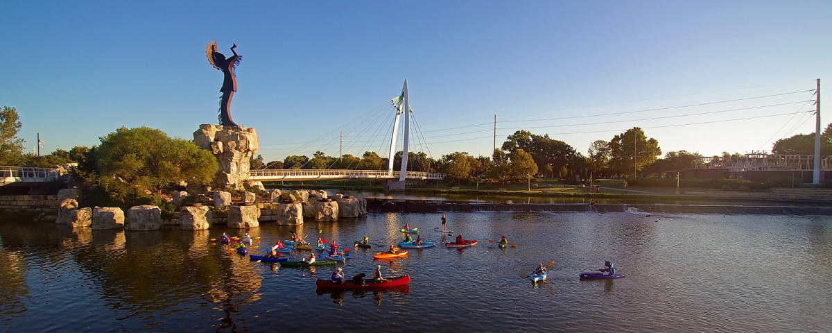 A group kayaks in the Arkansas River near the Keeper of the Plains statue
