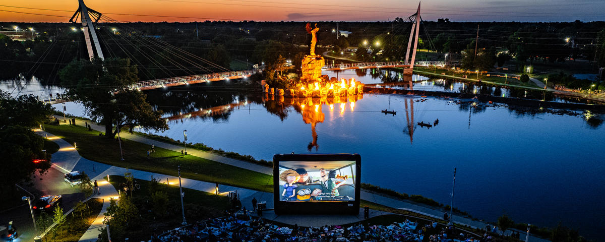 A crowd enjoys a movie screening during RiverFlix at the Wichita Foundation Amphitheater outside Exploration Place with the Keeper of the Plains fire pots burning in the background.