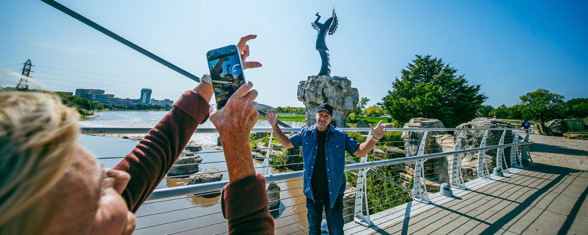 A man poses while another person takes a photo of him in front of the Keeper of the Plains on a sunny day in Wichita.