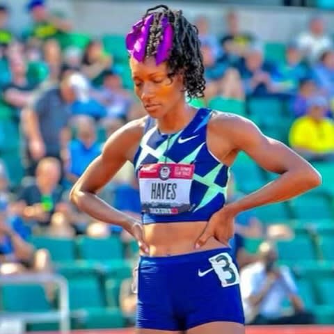 Elite sprinter Quanera Hayes stands on an outdoor track with her hands on her hips as she prepares to compete, with spectators seated in a stadium behind her.