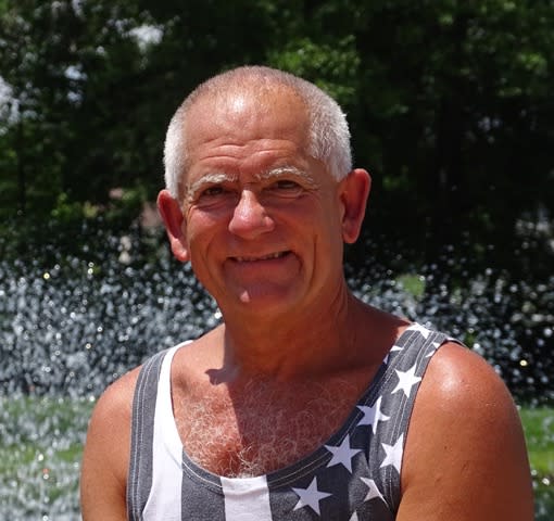 A tanned older man with white hair smiles at the camera. He's outside in front of some greenery and he's wearing a stars and stripes tank top