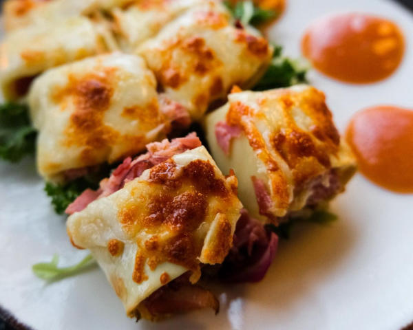 A close-up of VASO's smoked duck cannoli, topped with melted cheese, served with greens and sauce dollops on a white plate.