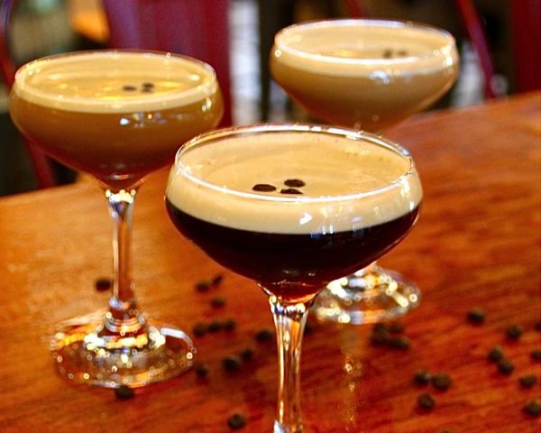 Three espresso martinis at Market Bar on a wood table with coffee beans, red chairs blurred in the background.