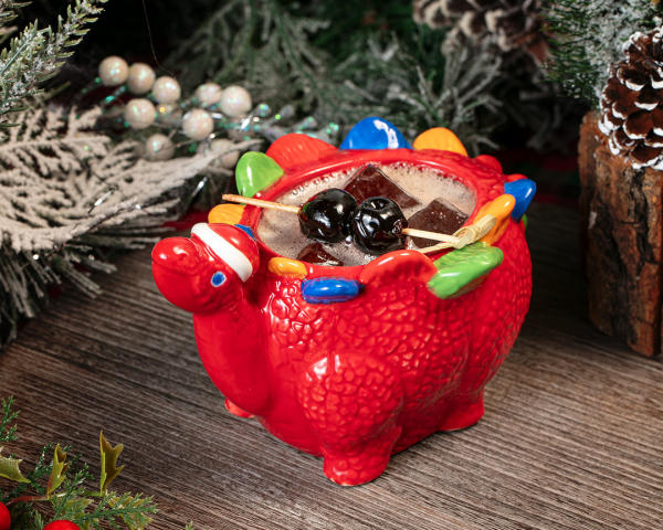 Holiday cocktail with cherries served in a red dinosaur mug, surrounded by festive greenery and pinecones.