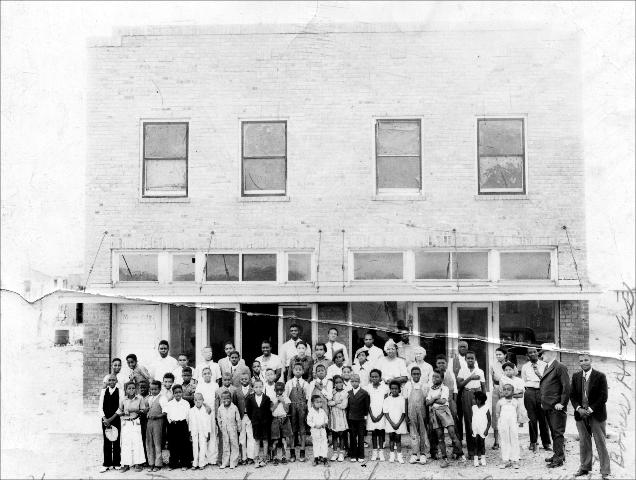 Bones Hooks and a group of people standing in front of "Pioneer Hall", which was located at 1511 NW Adams. The photograph is probably of Dogie Club members standing in front of Pioneer Hall. Bones Hooks is the man on the far right in the photo. The white man standing next to Bones is unidentified, but may have been Mr. Galloway.