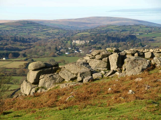Rolling countryside landscape with a foreground of large, weathered stone formations. In the distance, a stately manor lies amidst lush greenery.