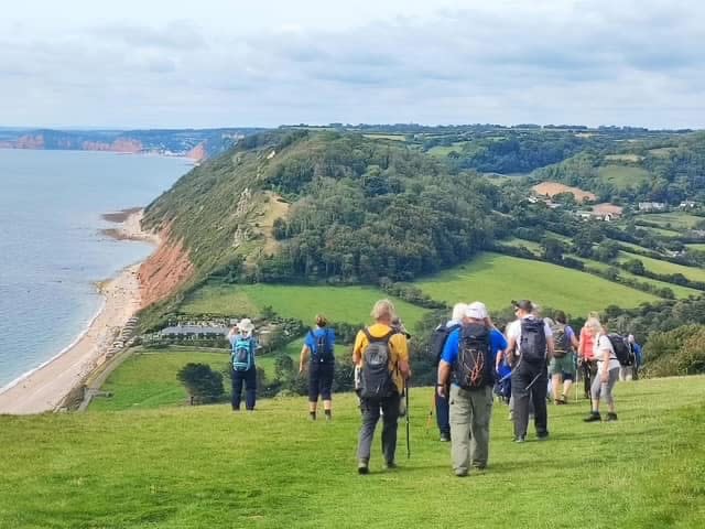 A group of hikers walks along a grassy cliffside trail overlooking the ocean and lush green hills. The sky is cloudy, conveying a serene, adventurous mood.