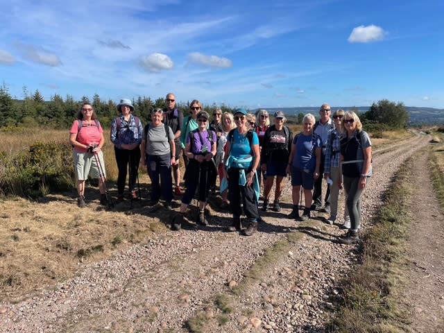 A group of 15 hikers stands on a gravel path in a rural landscape under a blue sky. They are wearing casual outdoor clothing, looking relaxed and happy.