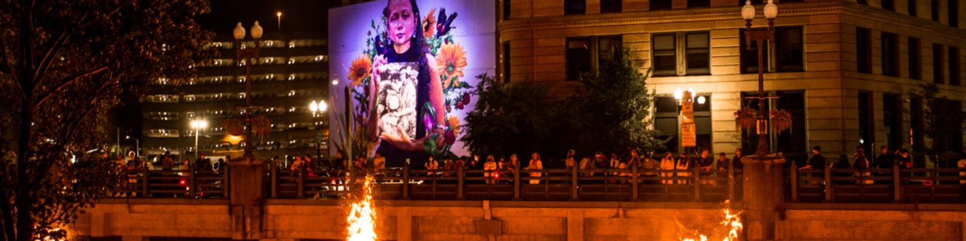 Braziers burn on the river and a mural of a young girl is illuminated in the background.