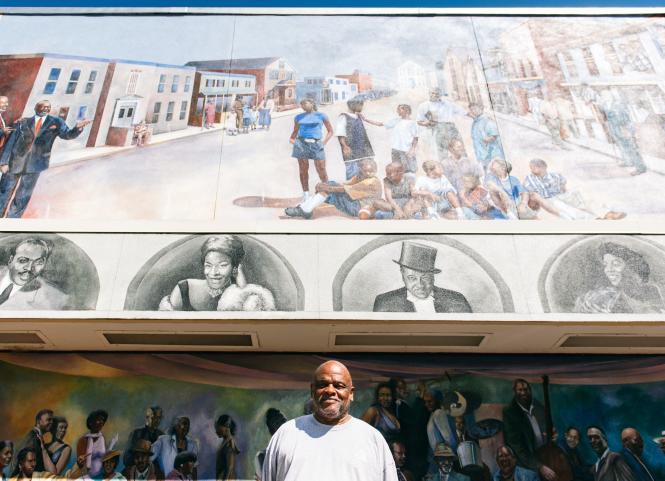 an African American man stands in front of a building with a mural behind him.