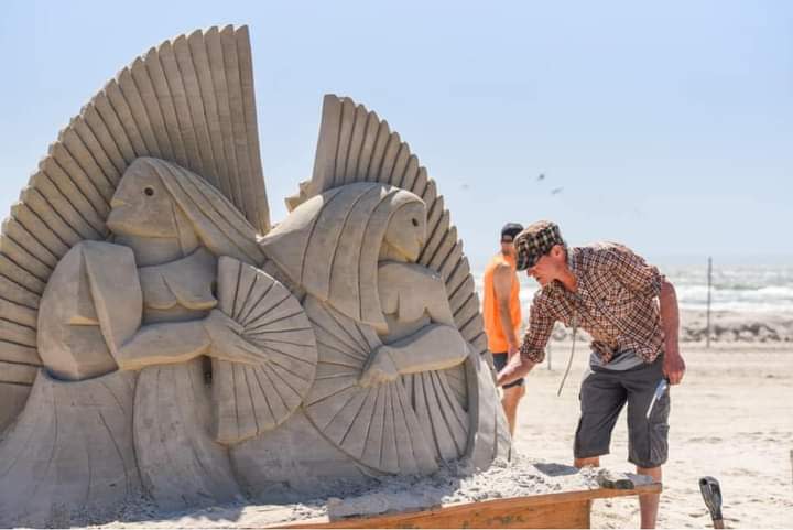 A man in shorts and a plaid shirt bends over a sand sculpture he's working on. The sculpture seem sto depict two figures holding fans, looking away from each other.