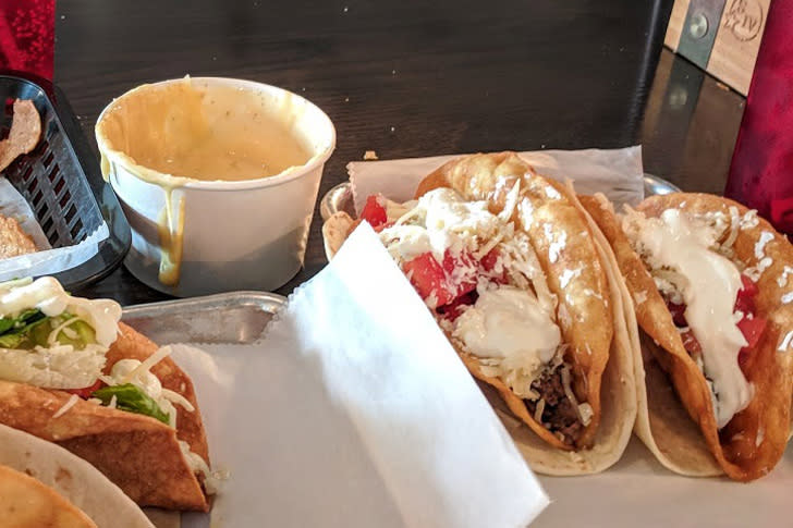 A table with two crunchy tacos topped with cheese and a white bowl of queso dip