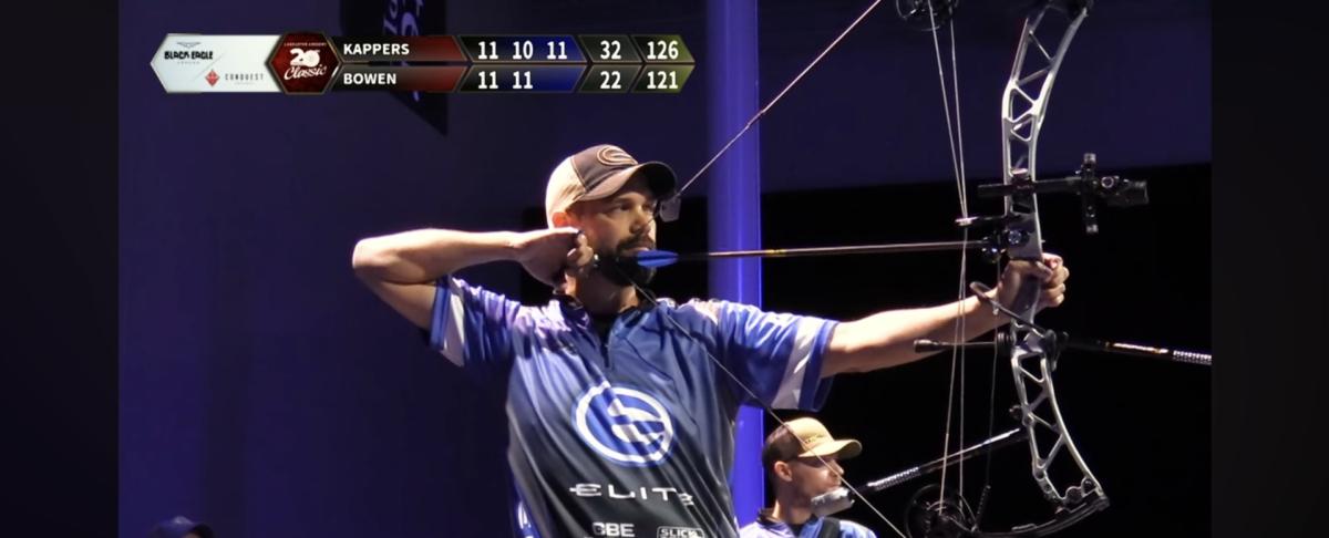 Richard Bowen draws a compound bow during an indoor archery competition. Wearing a blue Elite Archery team jersey and cap, Bowen takes aim while a live scoring graphic appears on-screen and another competitor waits in the background.