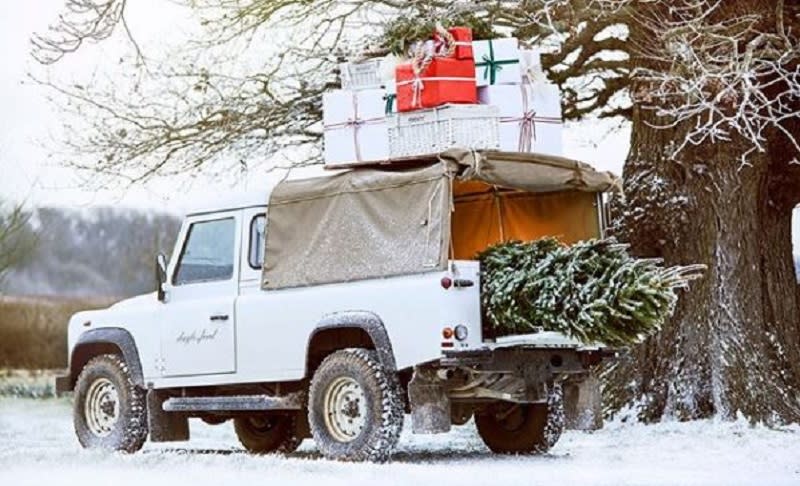 A Daylesford branded Landrover with Christmas tree sticking out the back and presents & hampers tied to the roof parked in snow covered grounds