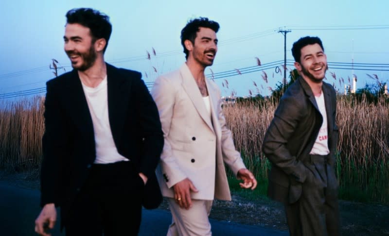 The three Jonas Brothers smile while wearing suits and walking through a field.