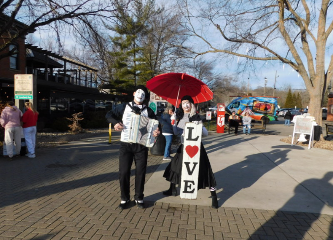 Image is of a male mime holding an accordion and a female mime holding an umbrella in one hand and a sign that says love in another.