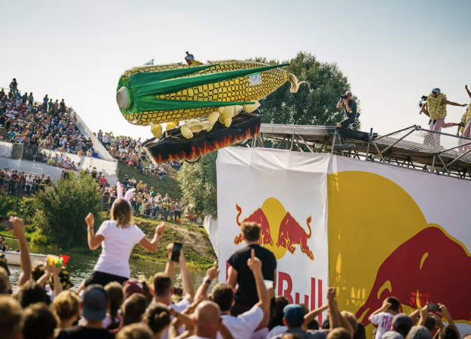 Image is of people in stands watching a giant corn on the cob looking plane, fly off a ramp with the Red Bull symbol.