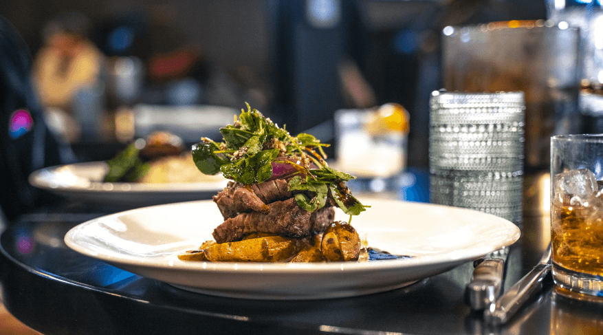 An elegant plated dish of steak and fresh greens, accompanied by a glass of whiskey, capturing the essence of a fine dining date night restaurant in Cheyenne.