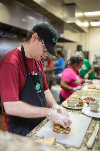 man in glasses, burgundy shirt and black apron prepares food in the kitchen