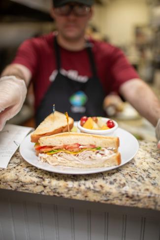 a plate with a sandwich and cup of fruit sitting on a counter with person in the background