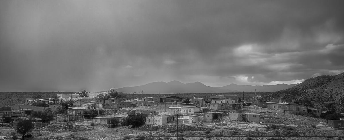 A small, remote village with simple houses sits under a cloudy, overcast sky. Mountains loom in the background.