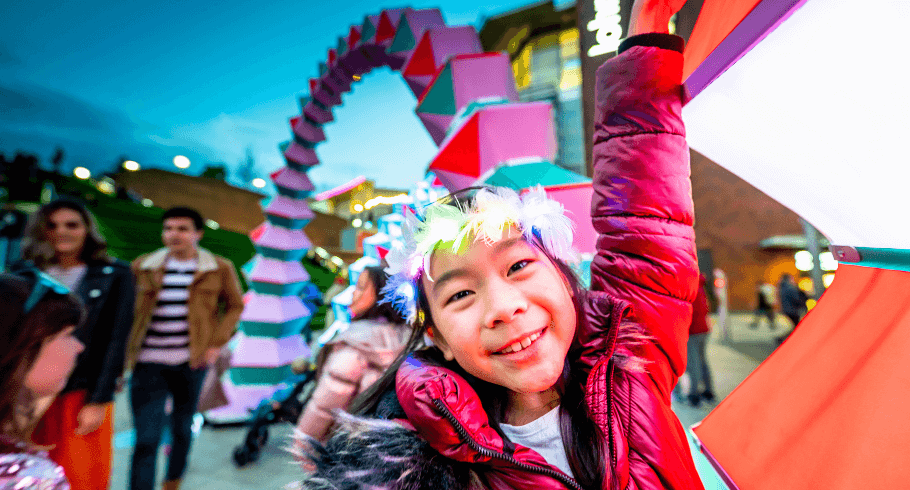 A young girl has a colourful, feathered headdress on and she smiles right at the camera as she touches an artwork