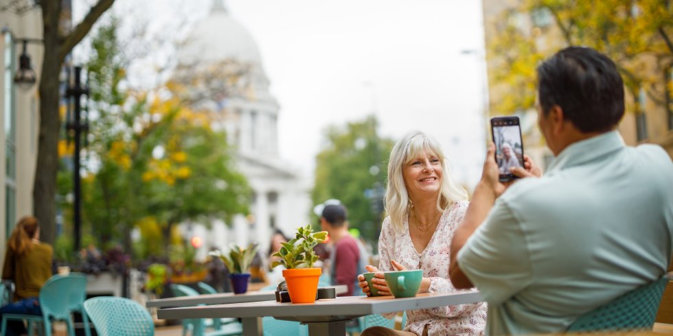 A man takes a picture of a woman with the Capitol building in the background as they enjoy sipping on coffee outside of Ancora coffee shop on King Street in downtown Madison