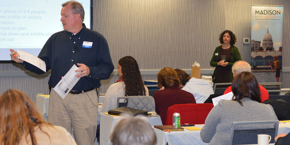 A man and woman give a PowerPoint presentation while others seated listen and take notes