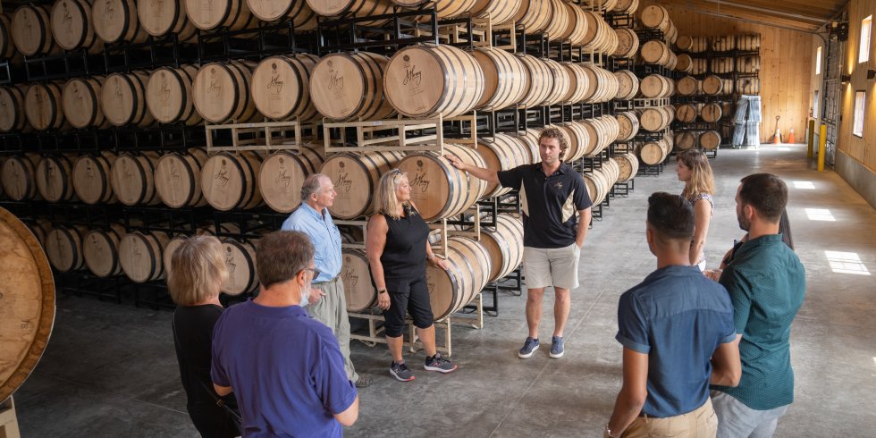 Eight people of varying ages and races gather around a tour guide, who is talking about the distillery process on a tour of J. Henry Bourbon Distillery. They are all standing in front of large racks of hundreds of barrels.