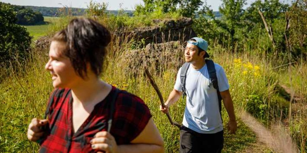 A woman and a man hiking through a grassy section of the Ice Age Trail on a sunny day. The woman is wearing a backpack and the man is carrying a walking stick.