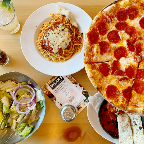 Aerial shot of pepperoni pizza, salad, pasta, spice shakers, and lemonade on wooden table