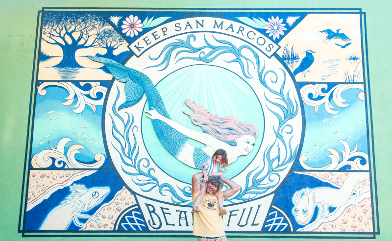 Father and daughter pose in front of a mural of a mermaid.