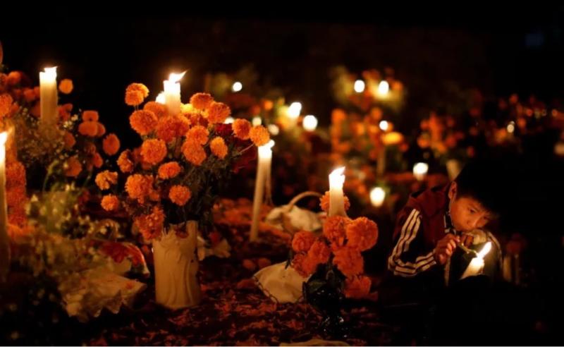 A young boy lights a candle among vibrant orange marigold flowers during a Día de los Muertos (Day of the Dead) celebration at night. The scene is illuminated by numerous candles, creating a warm and peaceful atmosphere in a cemetery, with offerings placed in remembrance of loved ones.