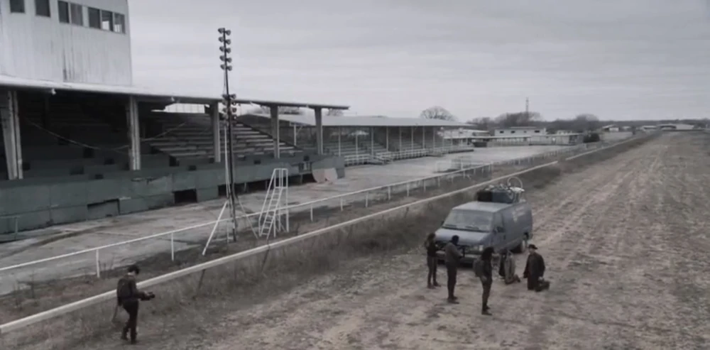 Fear the Walking Dead screengrab showing several masked figures holding two people at gunpoint. They are next to a parked car and standing on the deserted Ashburn Race Track