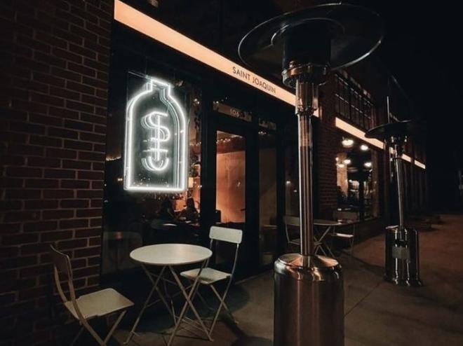 Neon sign hangs on bring wall of wine bar with table, chairs and heaters