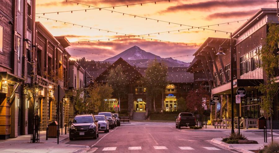 A charming street lined with shops and restaurants, illuminated by string lights at sunset, with a mountain backdrop. Cars are parked along the road.
