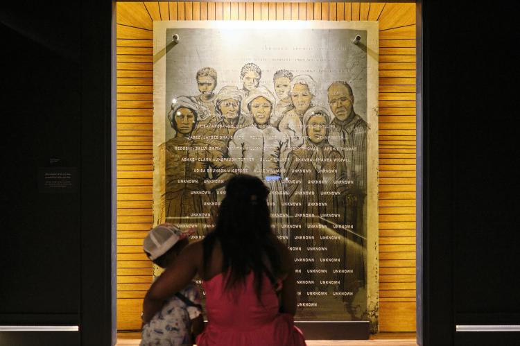A Black woman with long hair holds her child while they look at a photo collage of formerly enslaved residents of Africatown, Alabama